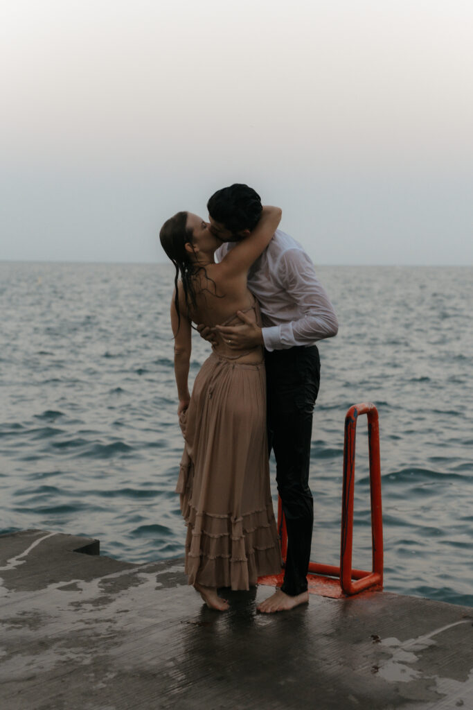 High-fashion embrace of a wet couple kissing passionately on a concrete pier after swimming in the ocean for their engagement photos.