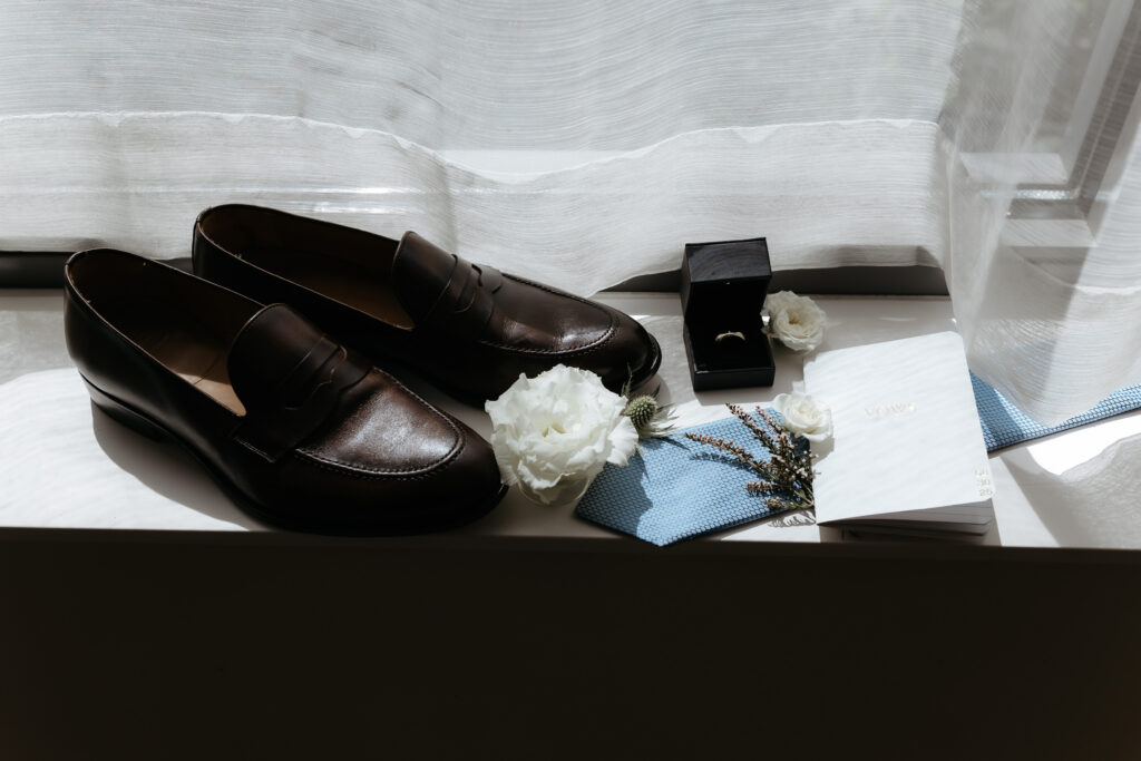 Editorial groom detail layout on a white windowsill featuring brown leather loafers, a blue tie, white flowers, a ring box, and a vow book.