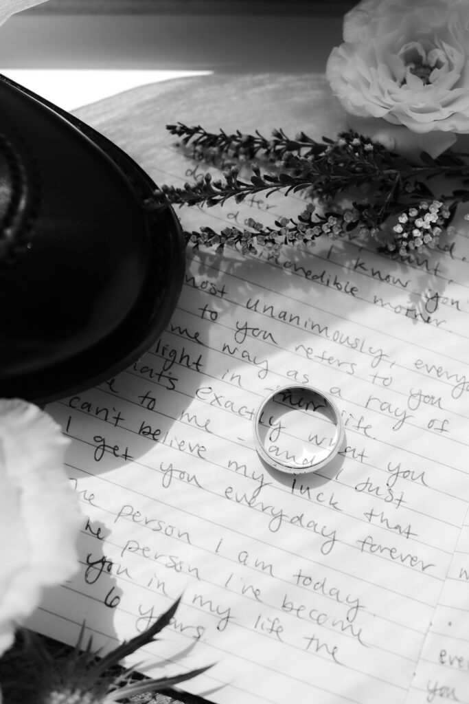 Black and white macro photo of a gold wedding band resting on a page of handwritten wedding vows next to a flower.