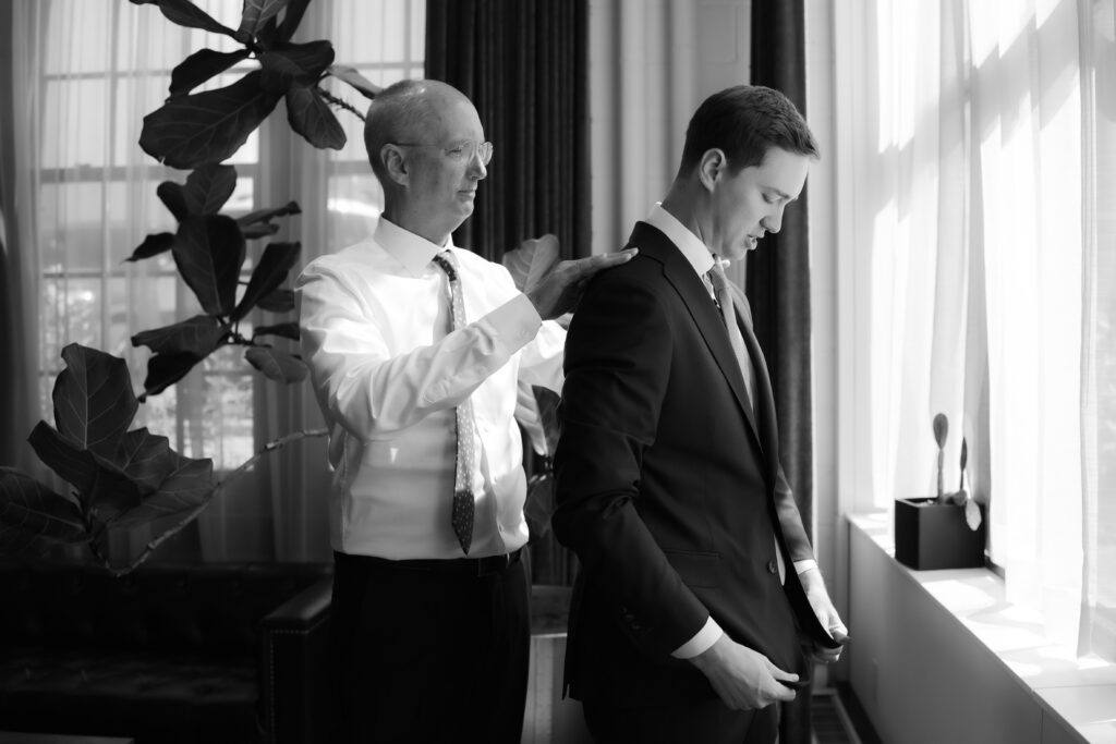 Black and white portrait of a father adjusting the shoulder of his son's wedding suit jacket by a bright window.