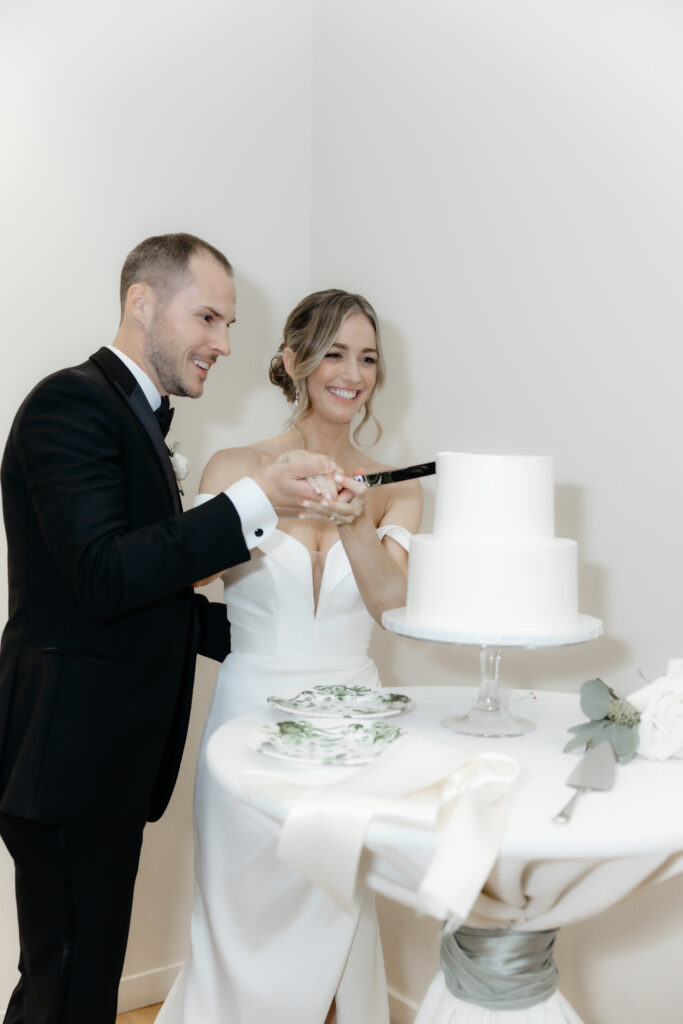 Smiling bride and groom holding a knife together to cut their white tiered wedding cake.