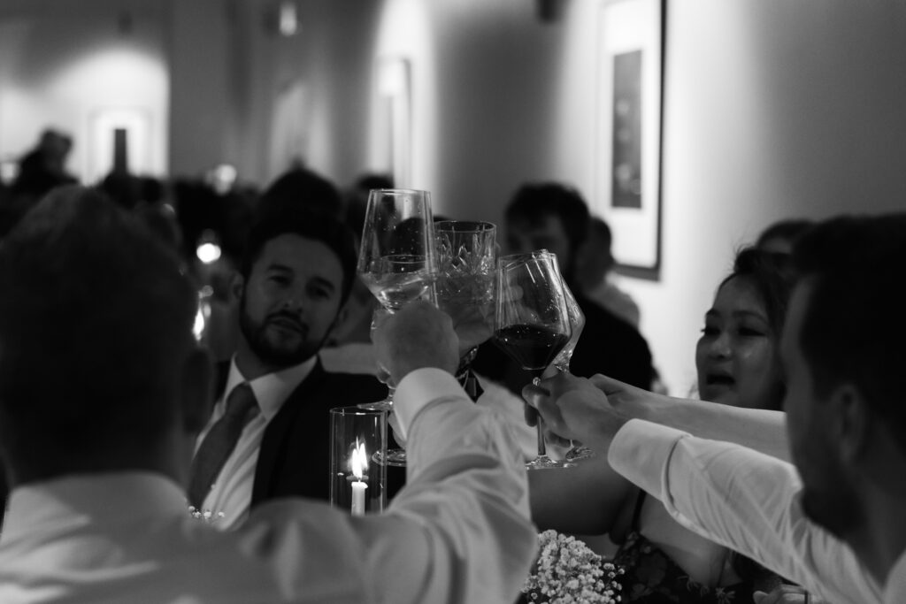 Black and white candid shot of wedding guests raising wine glasses for a dinner table toast.