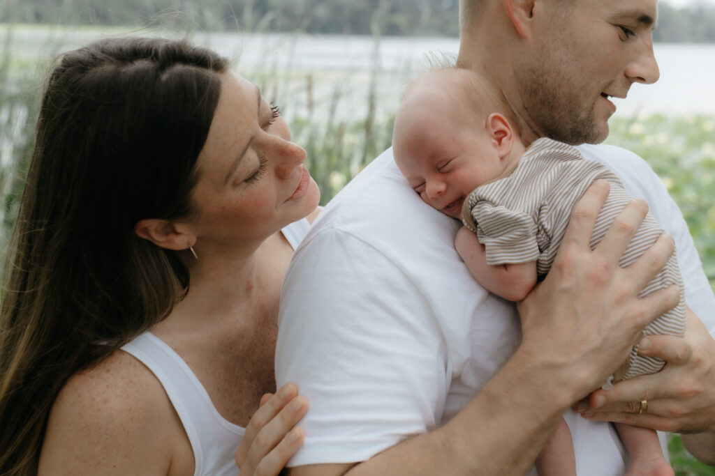 Mother smiling at her baby sleeping peacefully on her husband's shoulder outdoors.