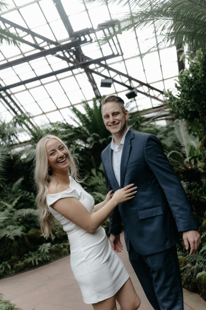 Happy engaged couple laughing during an editorial indoor session at a lush greenhouse with glass ceiling architecture.