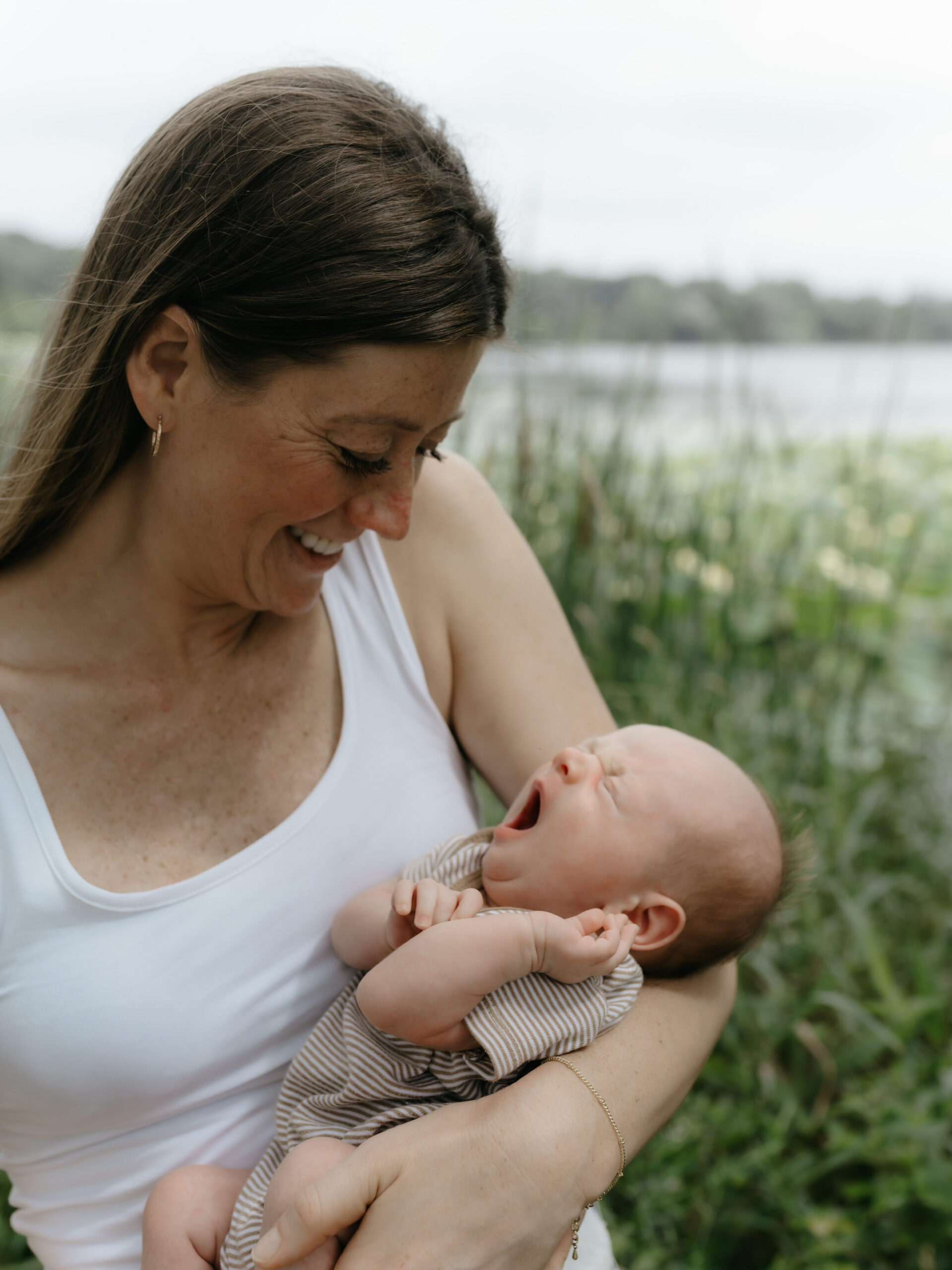 Mother holding her yawning newborn baby close to her chest in natural light.