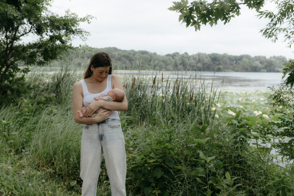 Mother holding her newborn baby in her arms while standing near tall park grass and a lake.