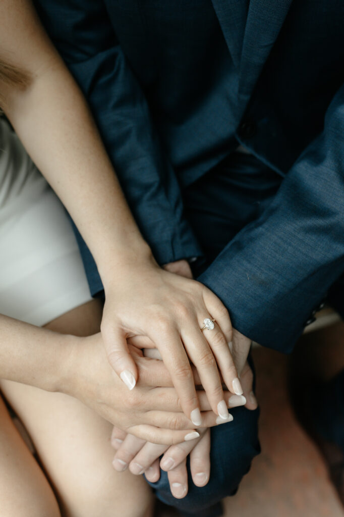 Close-up detail shot of an engaged couple's hands stacked on a navy suit lap showing an oval diamond engagement ring.