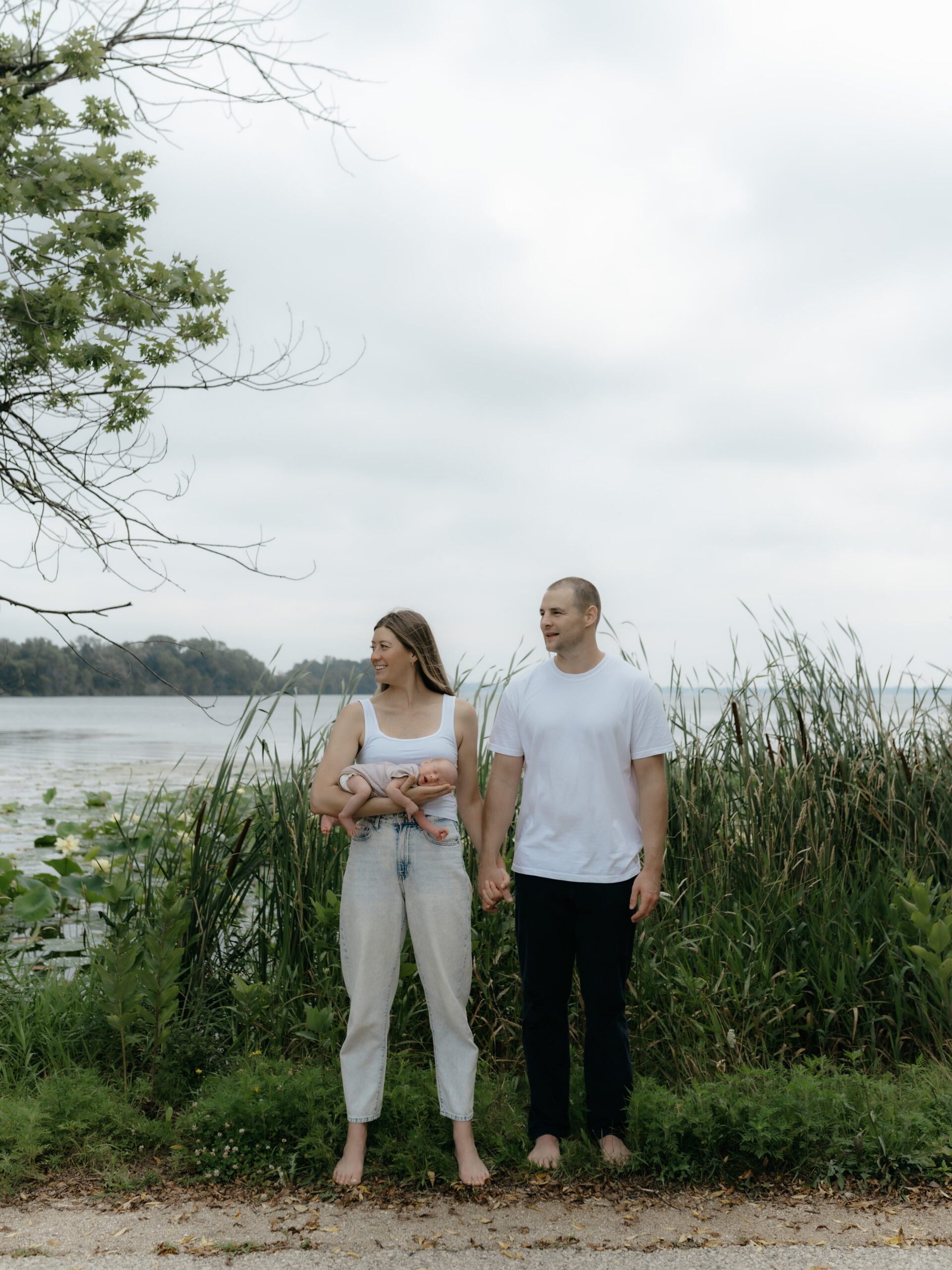 Parents holding hands while standing by a lake shore with their sleeping newborn.