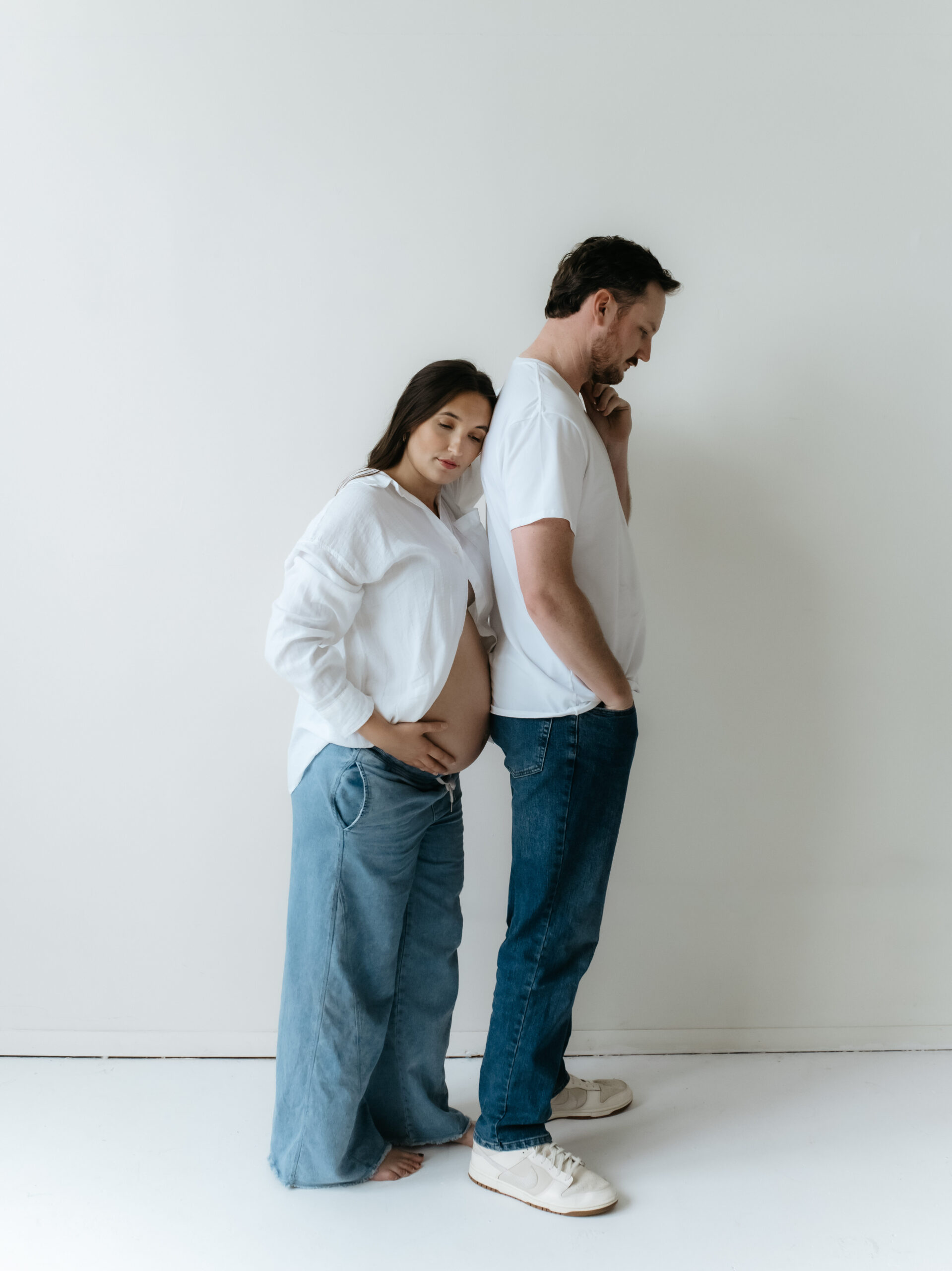 Full body shot of a couple standing back-to-back in matching casual white outfits against a seamless white backdrop.
