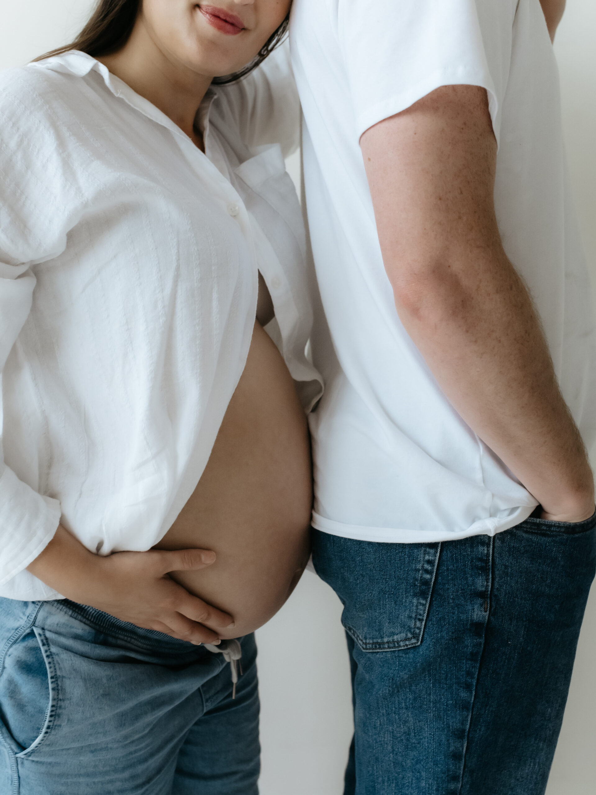 Close-up detail view of a pregnant belly and blue jeans held softly by hands in a white photo room.