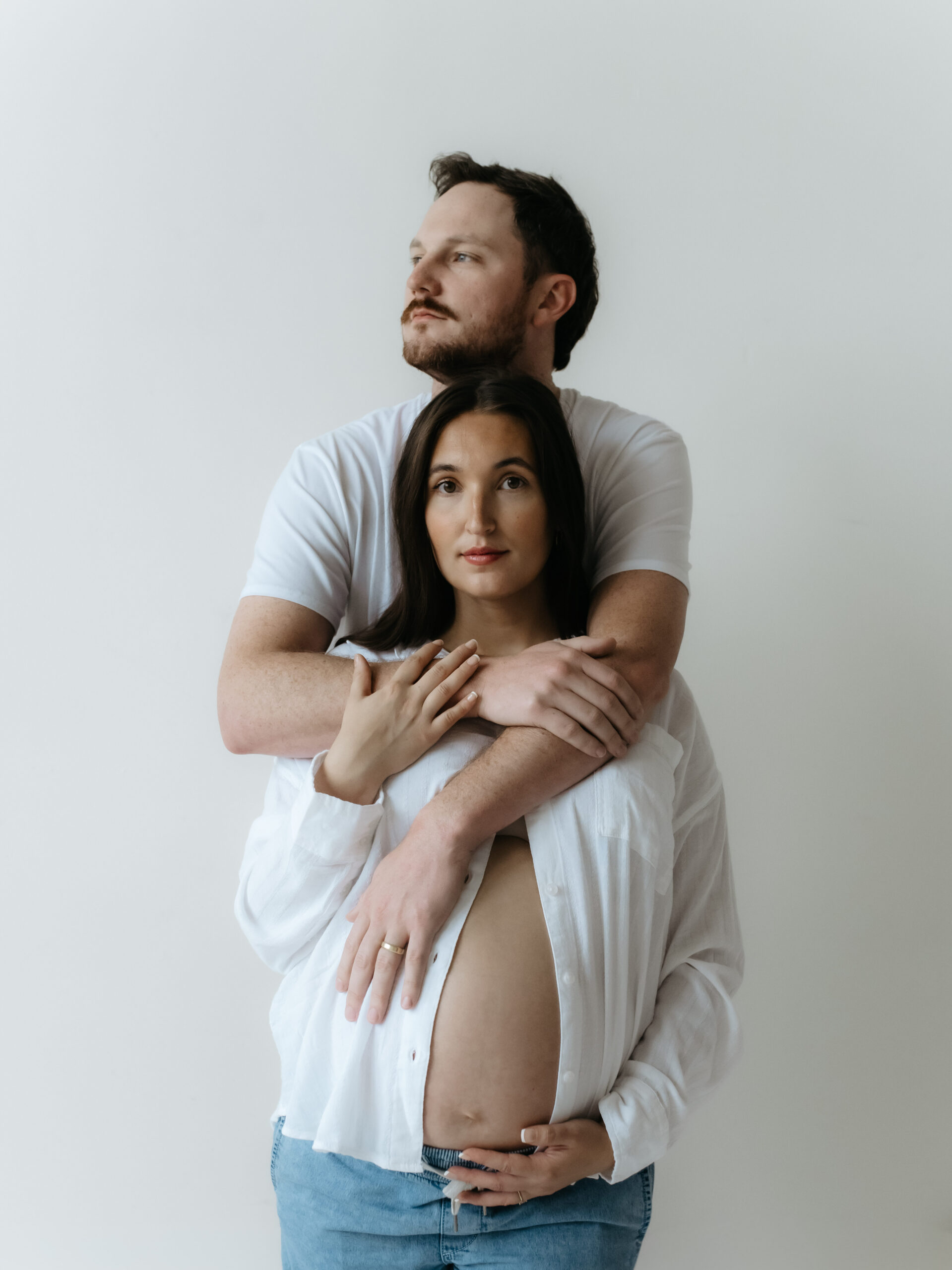 Man hugging his pregnant partner from behind against a plain white studio background while holding her bump.