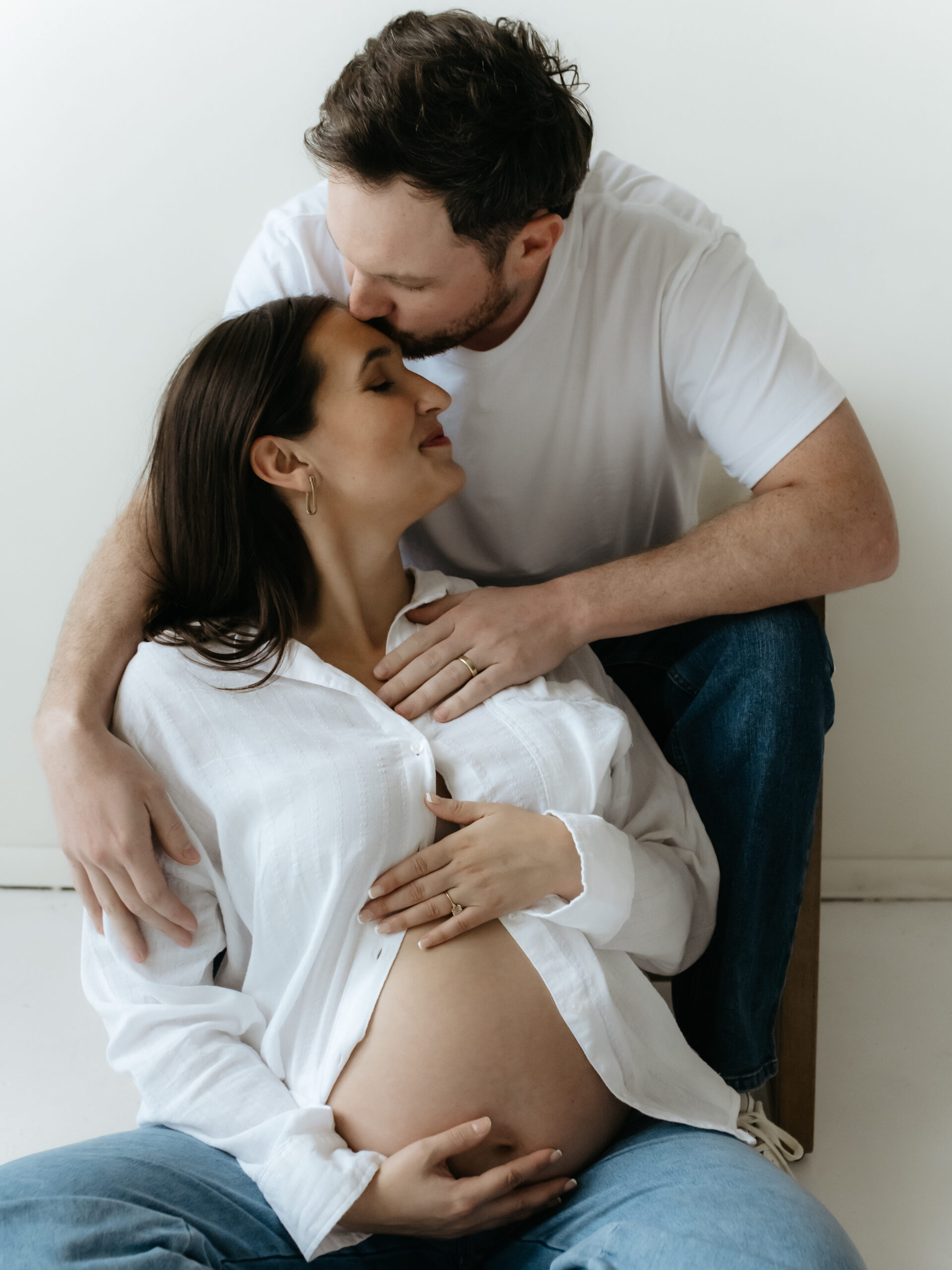 Husband kissing his seated pregnant wife on the forehead while both hold her baby bump.
