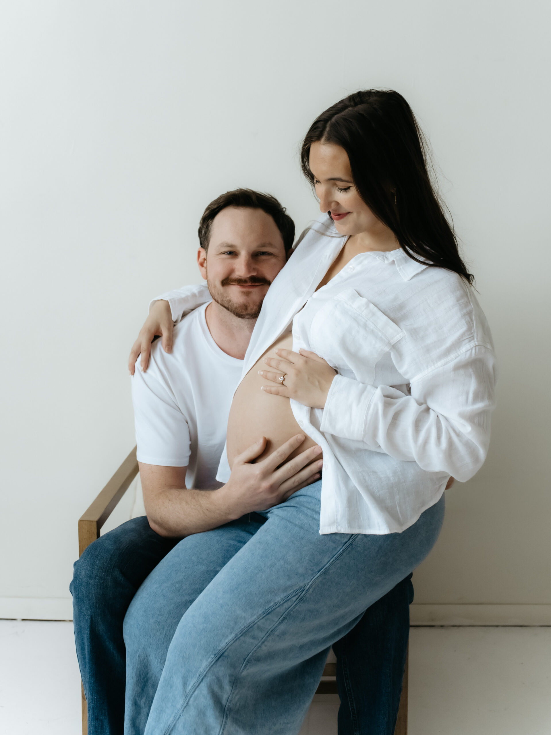 Pregnant woman sitting on her partner's lap in a studio chair smiling together in casual white outfits.