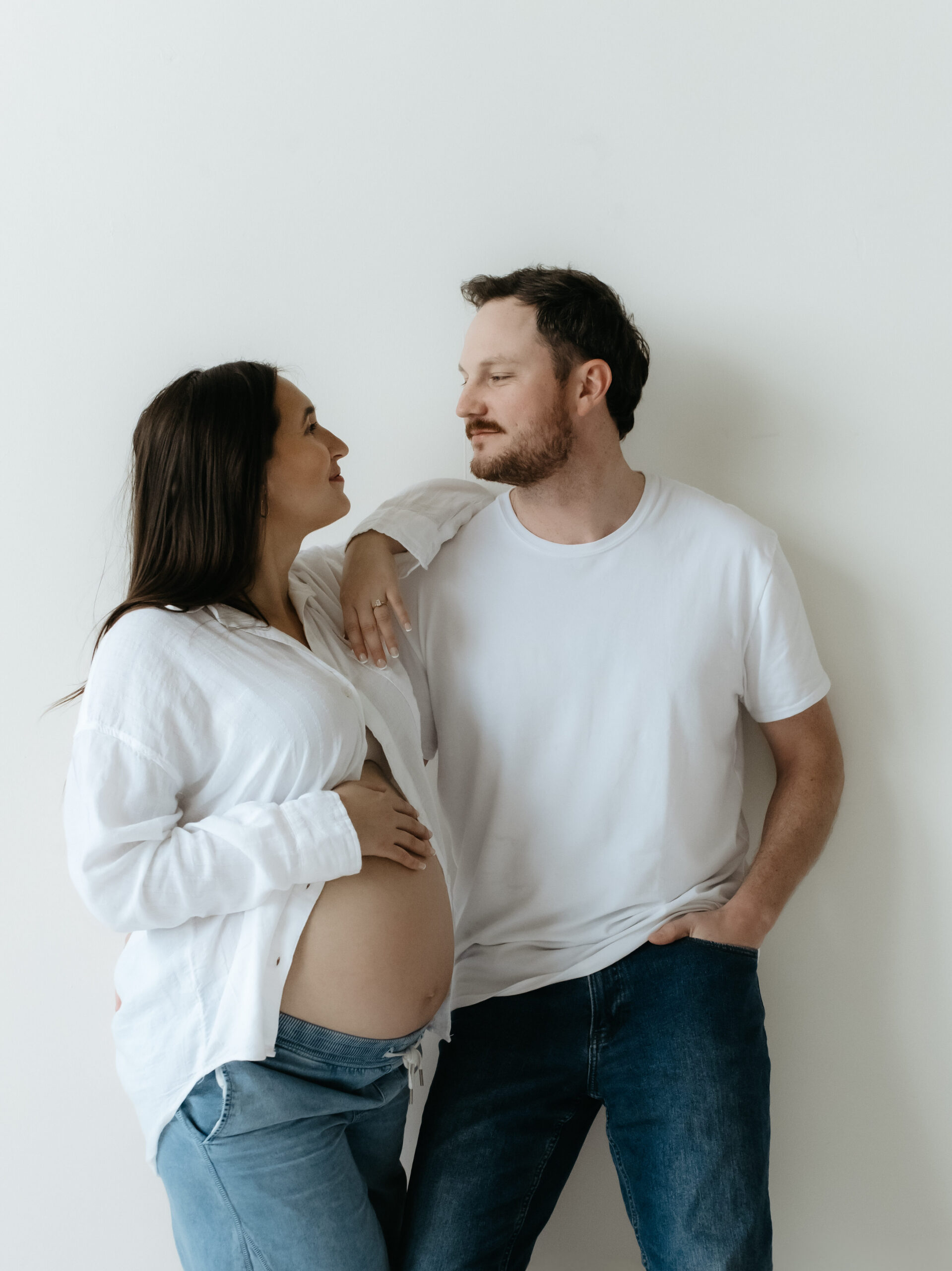 Couple in white tops and blue jeans leaning against a white studio wall looking at each other.