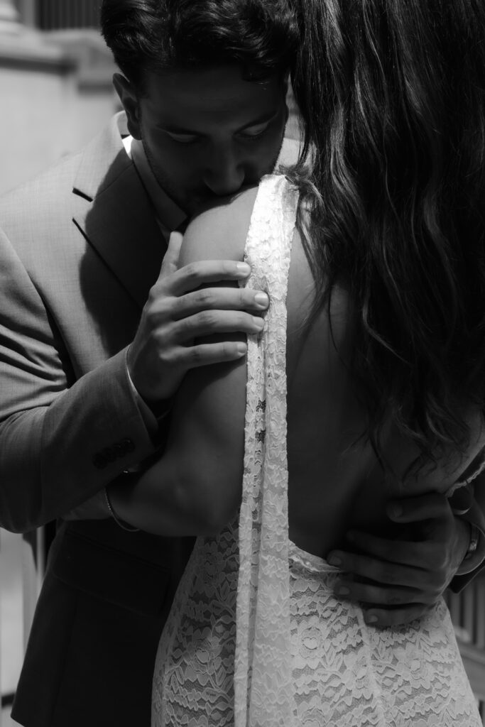 Close black and white portrait of a groom kissing his bride on the shoulder in strong side light.