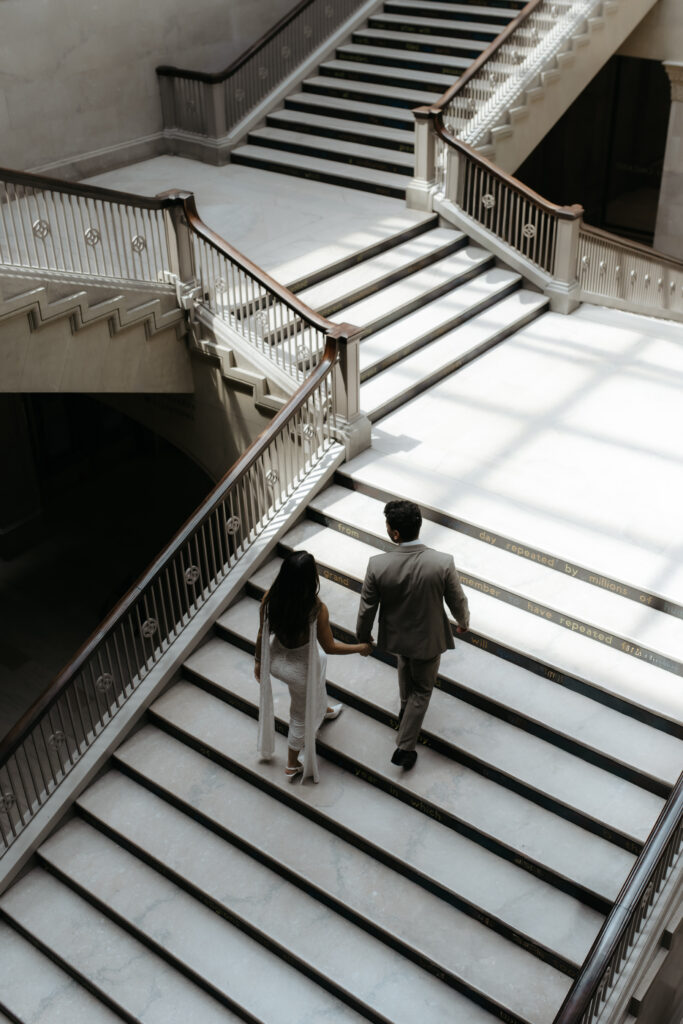 Back view of a couple holding hands as they walk up a wide marble staircase toward bright window light.