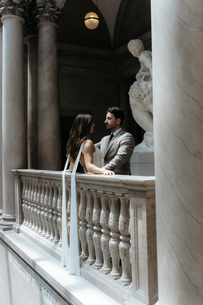 Couple leaning on a carved stone balcony railing next to tall marble pillars in a museum hall.