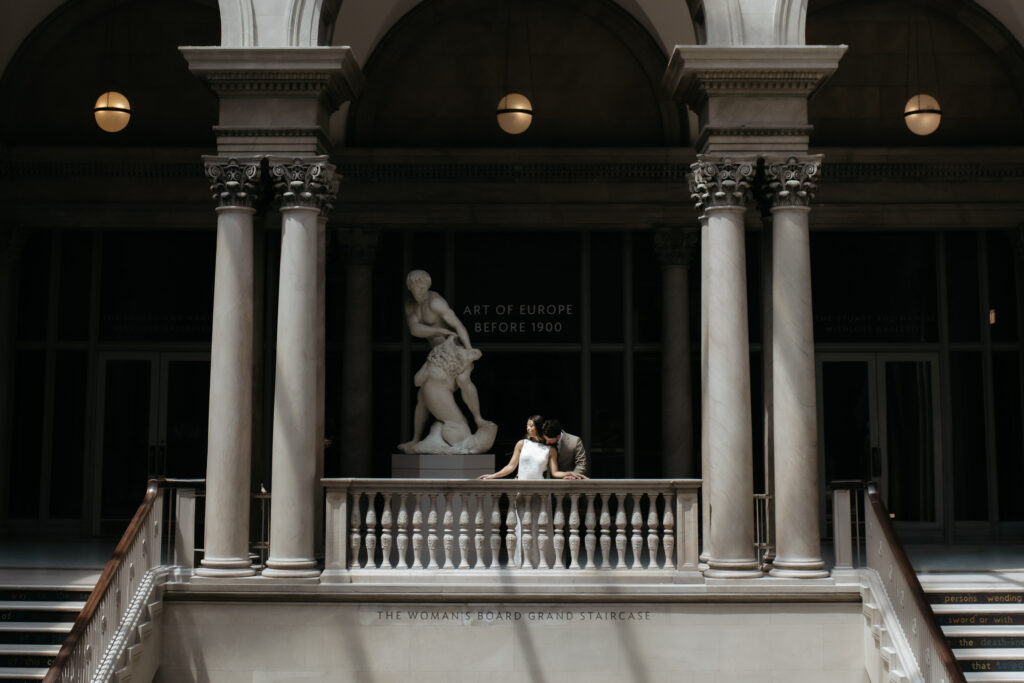 Wide view of a couple standing on a stone balcony framed by large pillars and statues under a skylight pool of light.