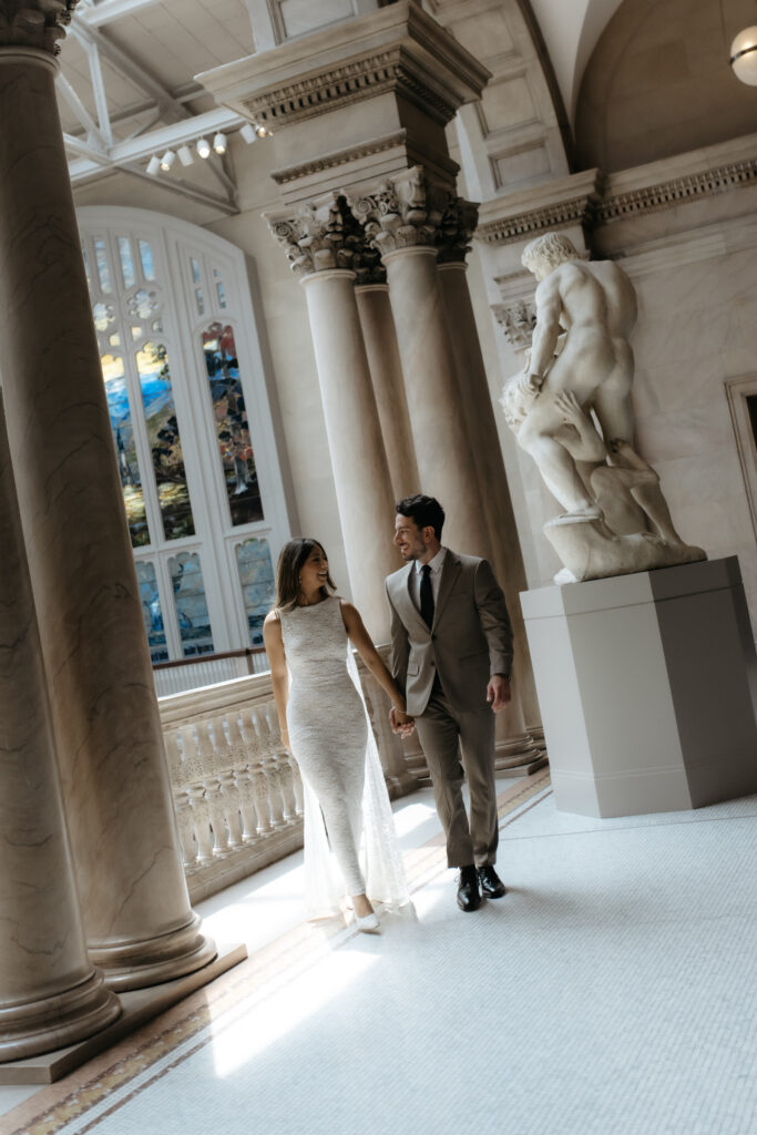 Bride in a lace dress and groom walking along a museum balcony lit by a overhead beam of sunlight.