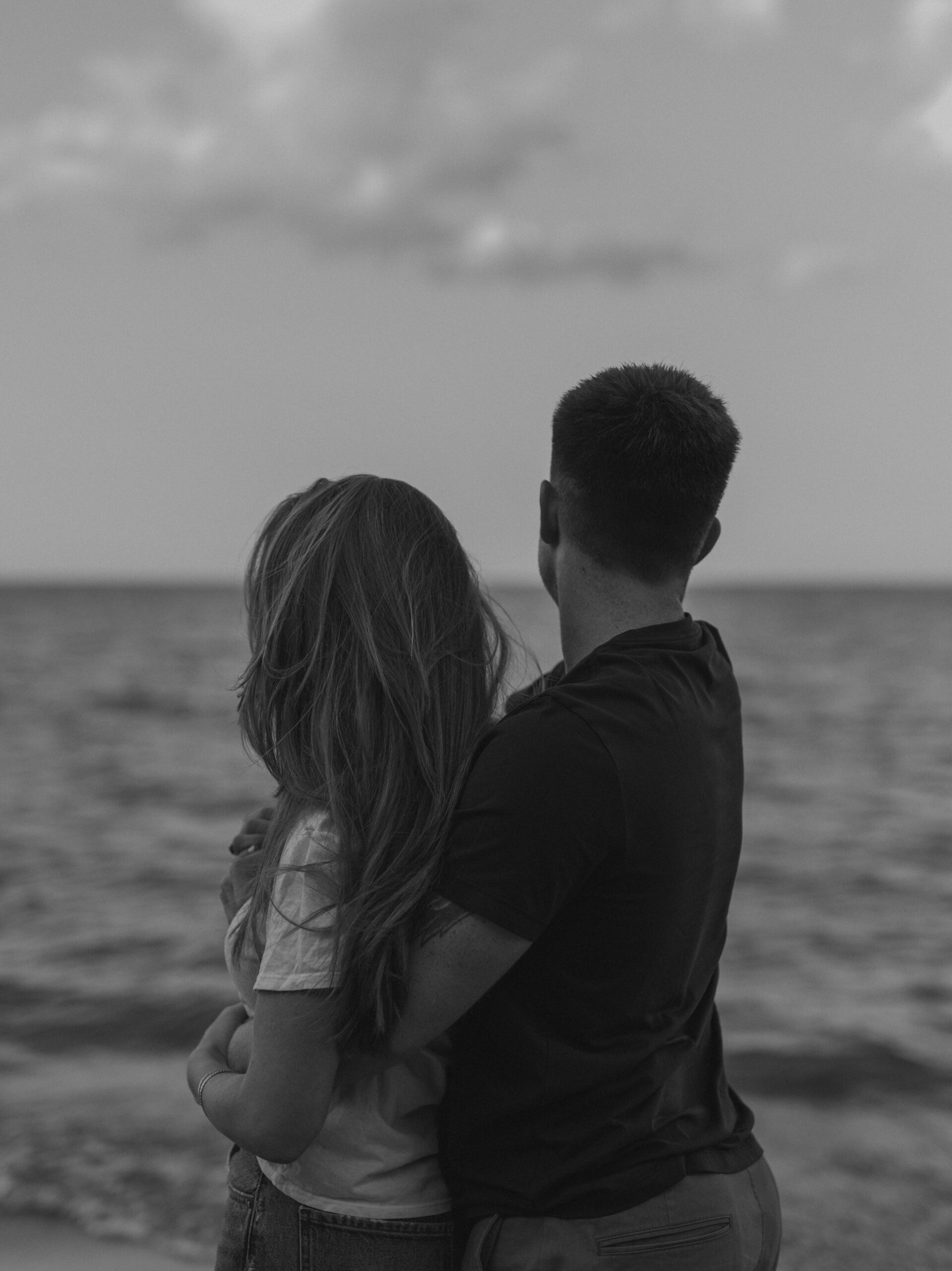 Black and white shot of a couple standing by the water looking out at the calm sea together.