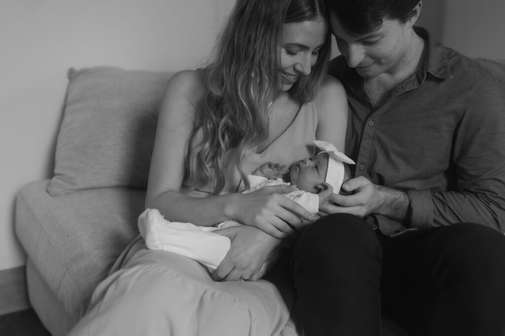 Black and white close-up of a mother and father holding their sleeping baby girl on a cozy couch.