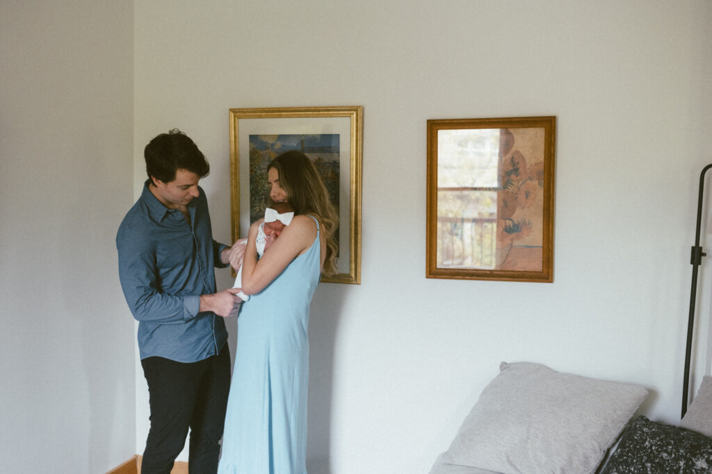 Parents standing together in a light-filled living room cradling their newborn baby in a blue dress.