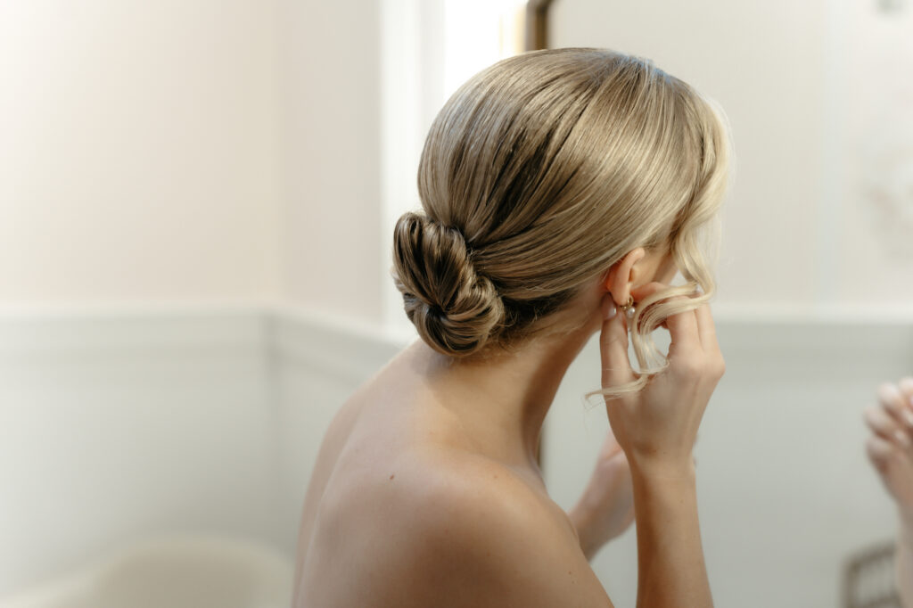 Close-up of a bride putting on a pearl earring showing her sleek low hair bun.