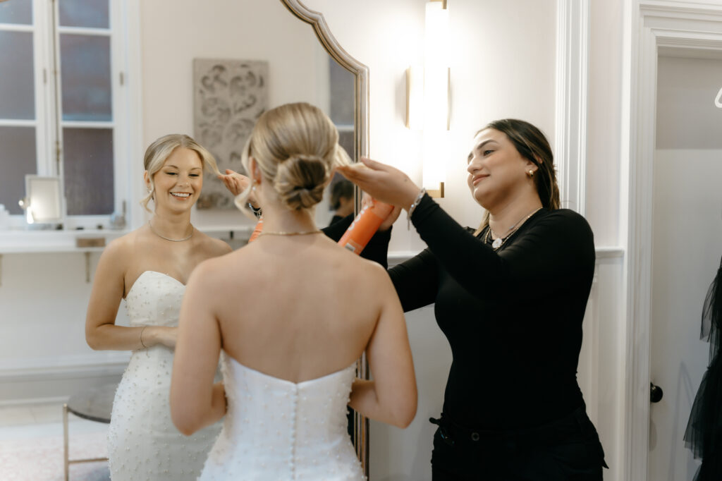 Hairstylist applying hairspray to a bride in a pearl-embellished gown reflecting in a gold mirror.