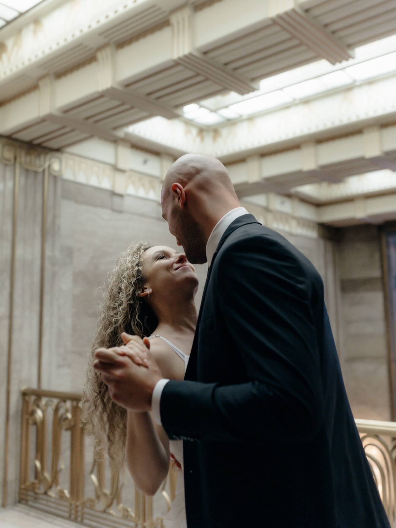A bride smiling up at her groom while slow dancing under a vaulted glass ceiling inside an indoor historic venue.