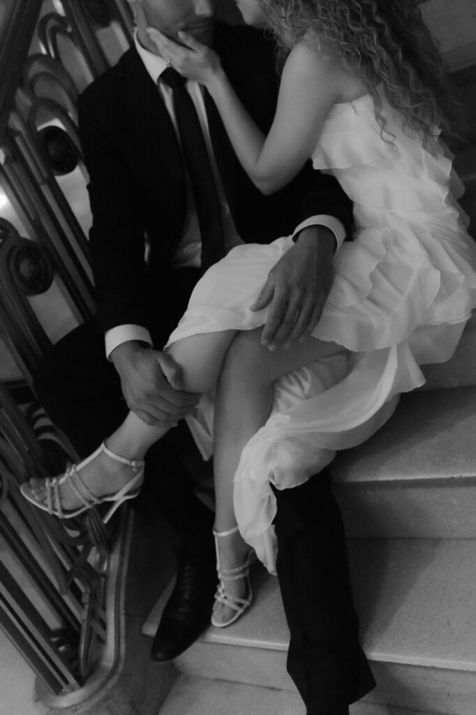 Black and white close-up shot of a bride sitting on her groom's lap on a marble staircase.