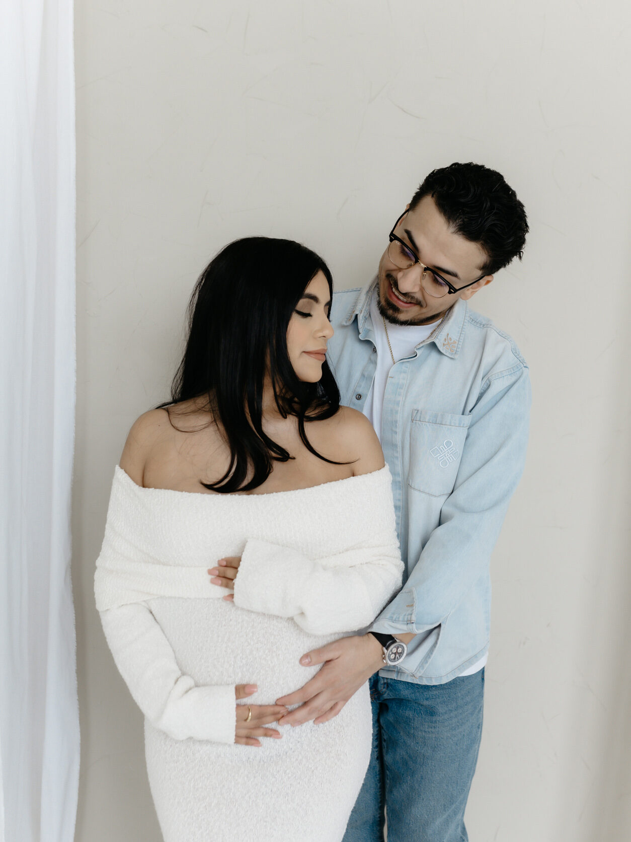 An affectionate husband hugging his pregnant wife wearing a cream off-the-shoulder knit dress during an indoor studio maternity session.