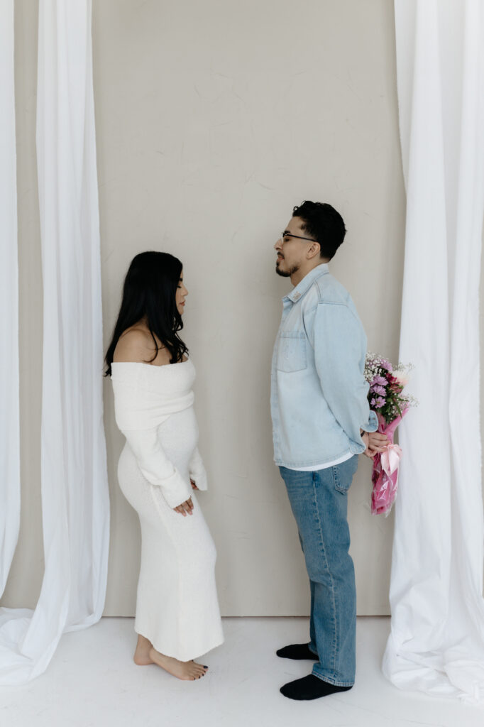 A husband hiding a colorful floral bouquet behind his back while standing opposite his pregnant wife on a white studio floor.
