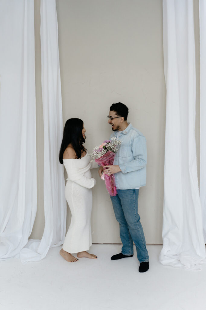 A husband surprising his pregnant wife with a fresh flower bouquet inside a bright white studio setting.