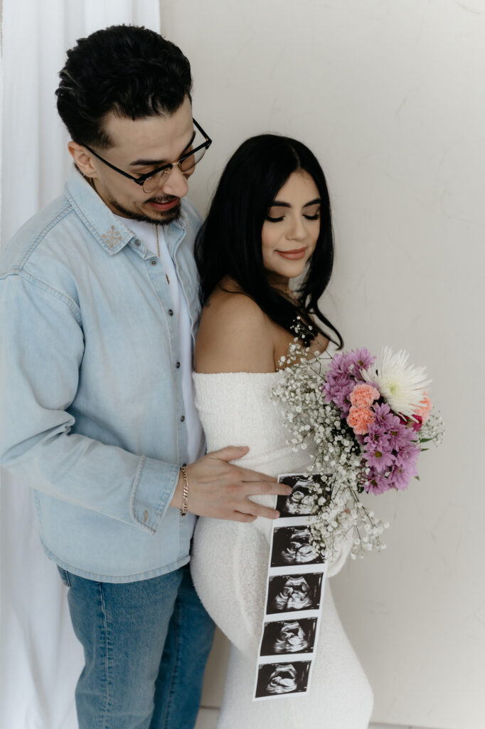 A close-up portrait of an expecting couple holding a flower bouquet and a strip of ultrasound pictures against a pregnant belly.