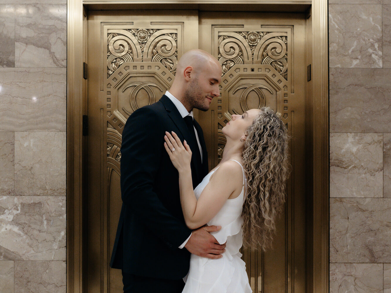 An elegant couple in a tux and white tiered dress embracing in front of ornate brass elevator doors inside a historic marble lobby.
