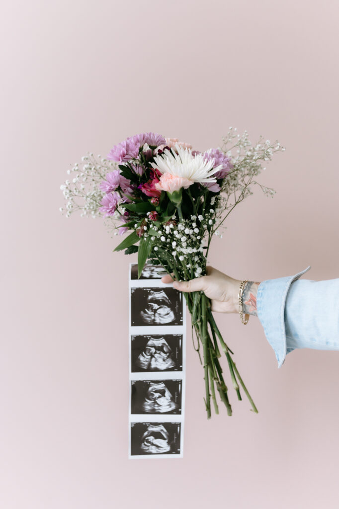 Detail shot of a hand holding a colorful bouquet of flowers attached to a long strip of baby ultrasound photos against a pink backdrop.