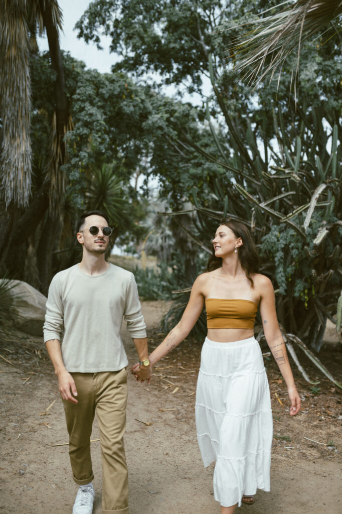 A cool couple holding hands and walking down a sandy desert path wearing wire-rim sunglasses and casual earth-tone outfits.