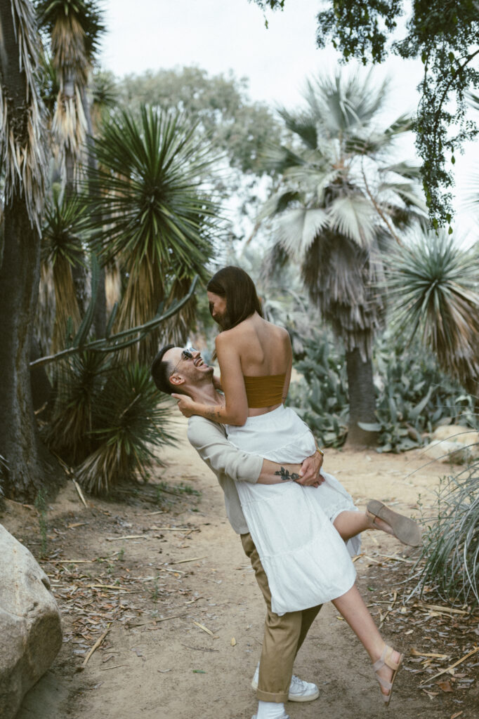 A man lifting his partner up in his arms and laughing while surrounded by tropical palm trees.