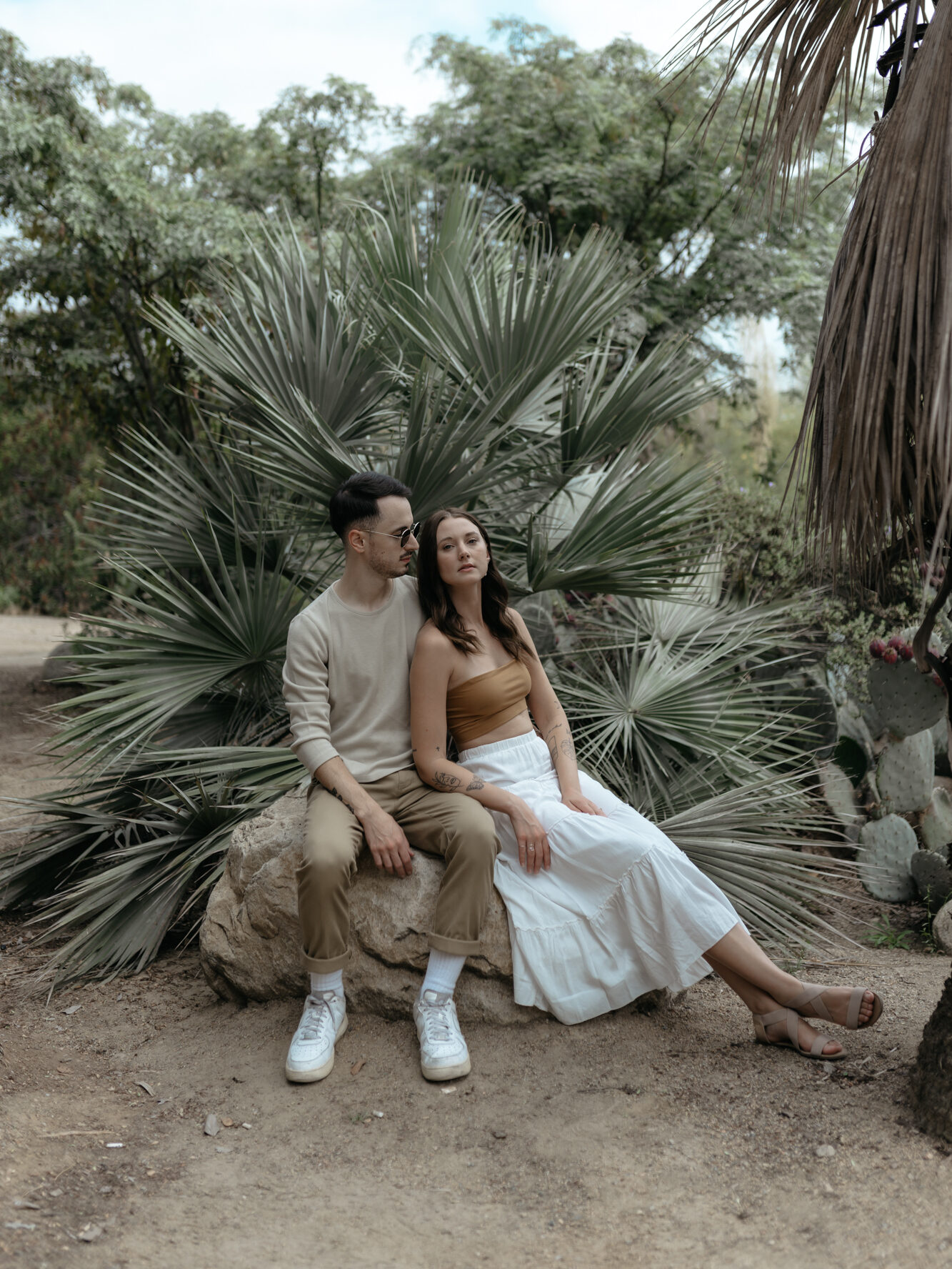 A stylish couple sitting together on a boulder in front of palm foliage in casual earthy outfits.