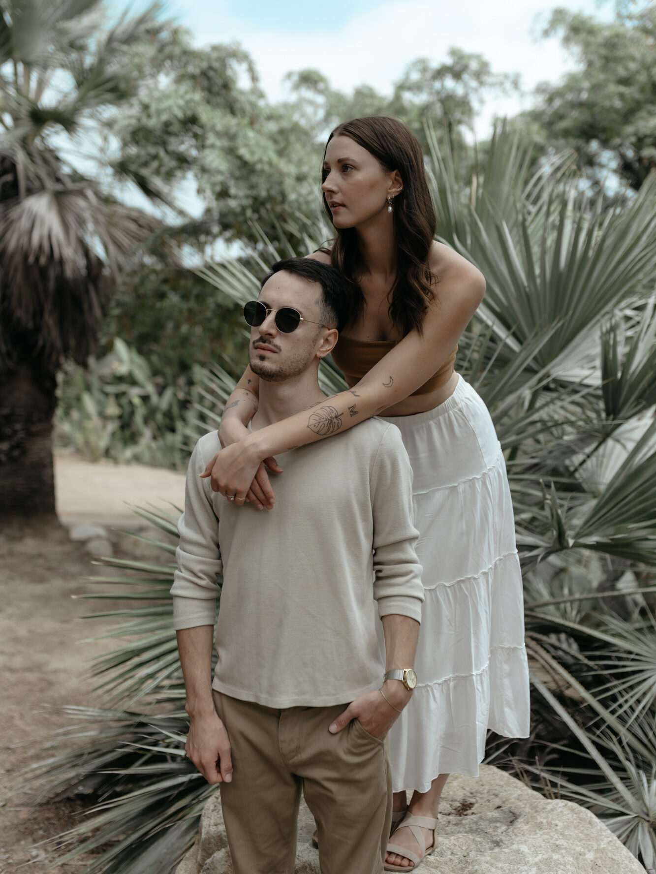 Editorial portrait of a woman leaning over her partner's shoulders while standing together in front of fan palm trees.