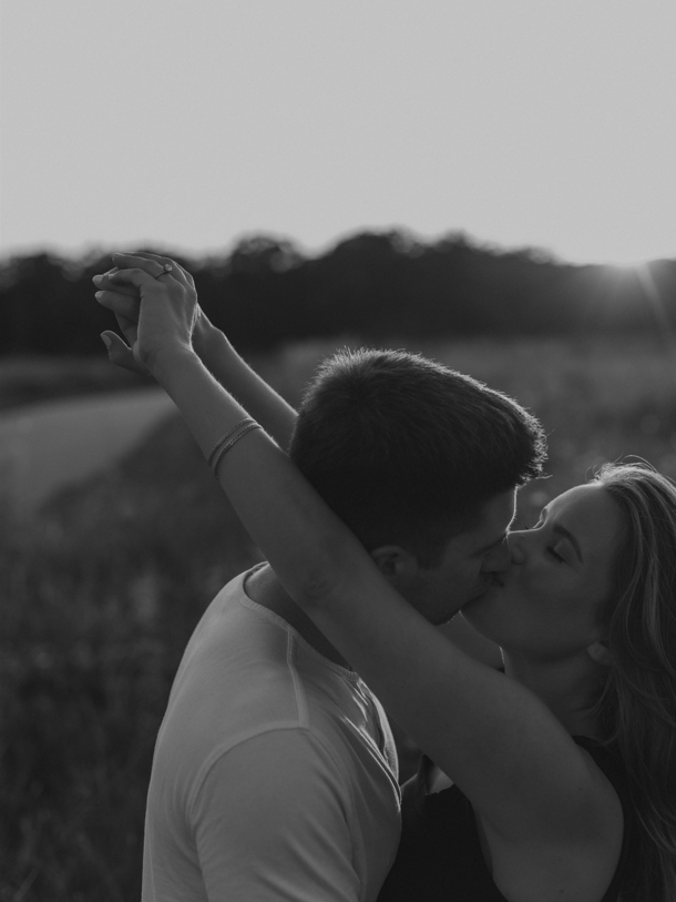 Black and white portrait of a couple kissing outdoors with their arms raised against a soft sunset sky.