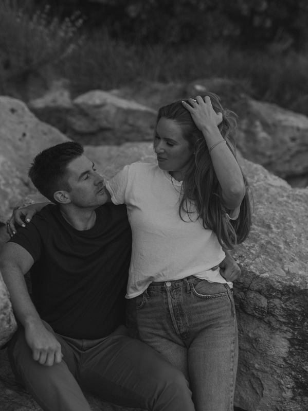 Black and white image of a couple sitting on large rocks outdoors looking into each other's eyes.