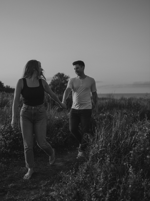Black and white portrait of a couple walking hand-in-hand along a grassy path near the shore.