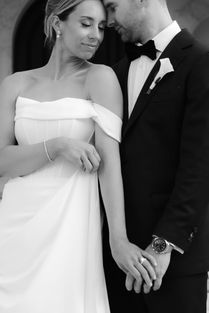 Black and white close up of a bride leaning her head against her groom while holding hands.