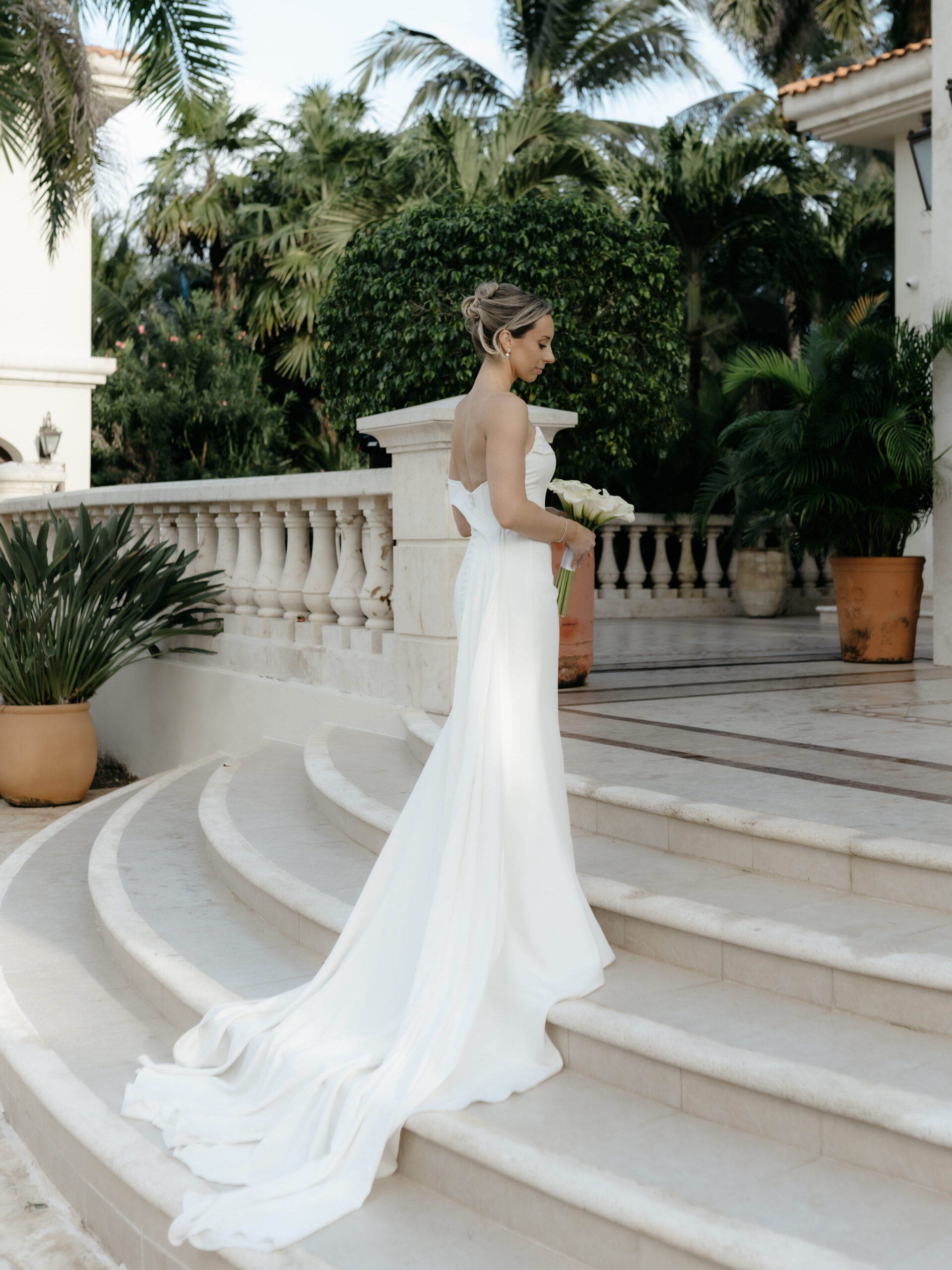 Bride standing alone on curved white stone villa steps holding a calla lily bouquet.