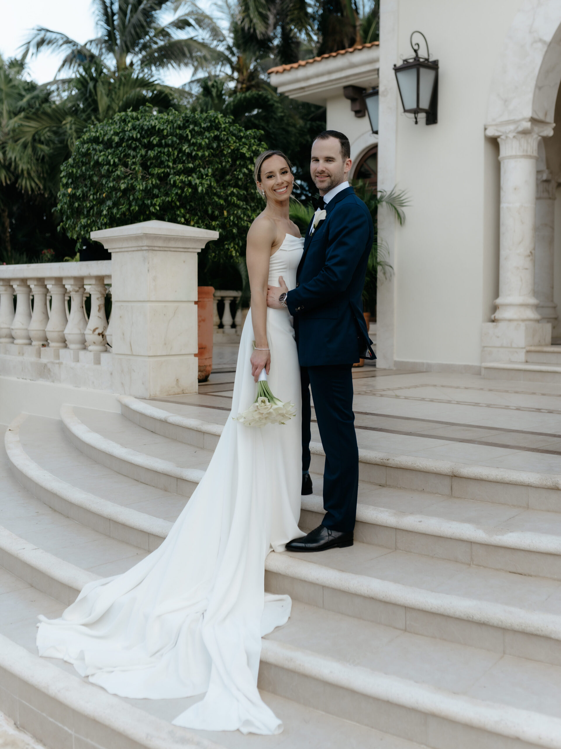 Bride and groom smiling together on outdoor stone steps surrounded by tropical palm trees.