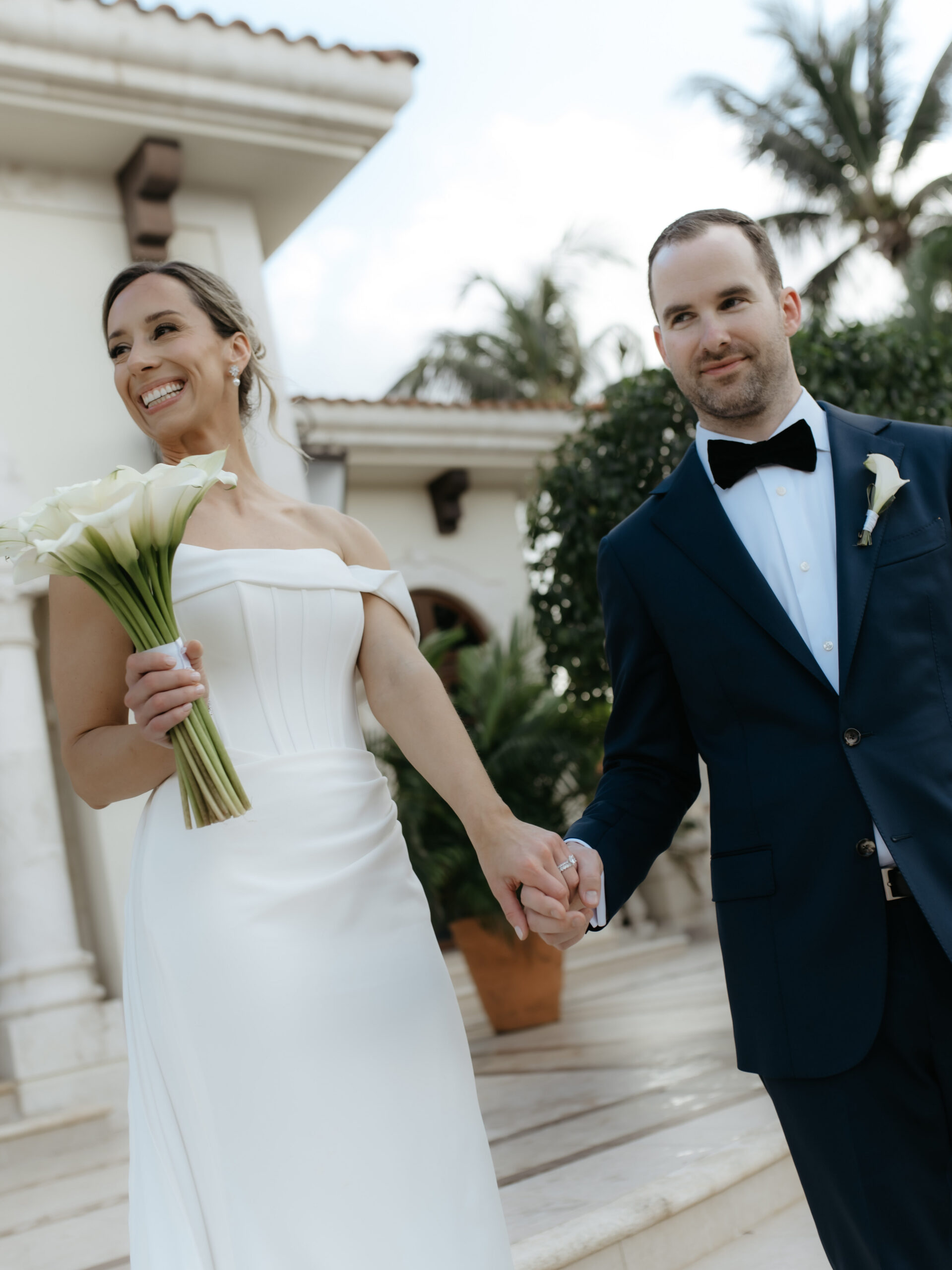 Bride and groom holding hands and laughing while walking down a villa path.