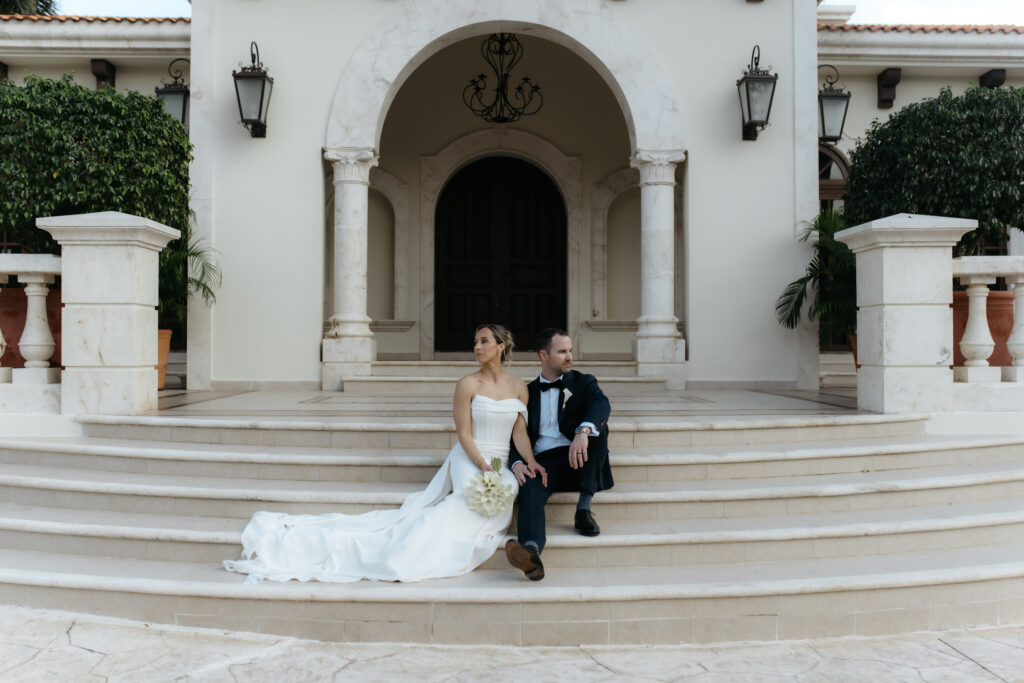 Couple sitting together on white stone steps in front of a grand arched entryway.