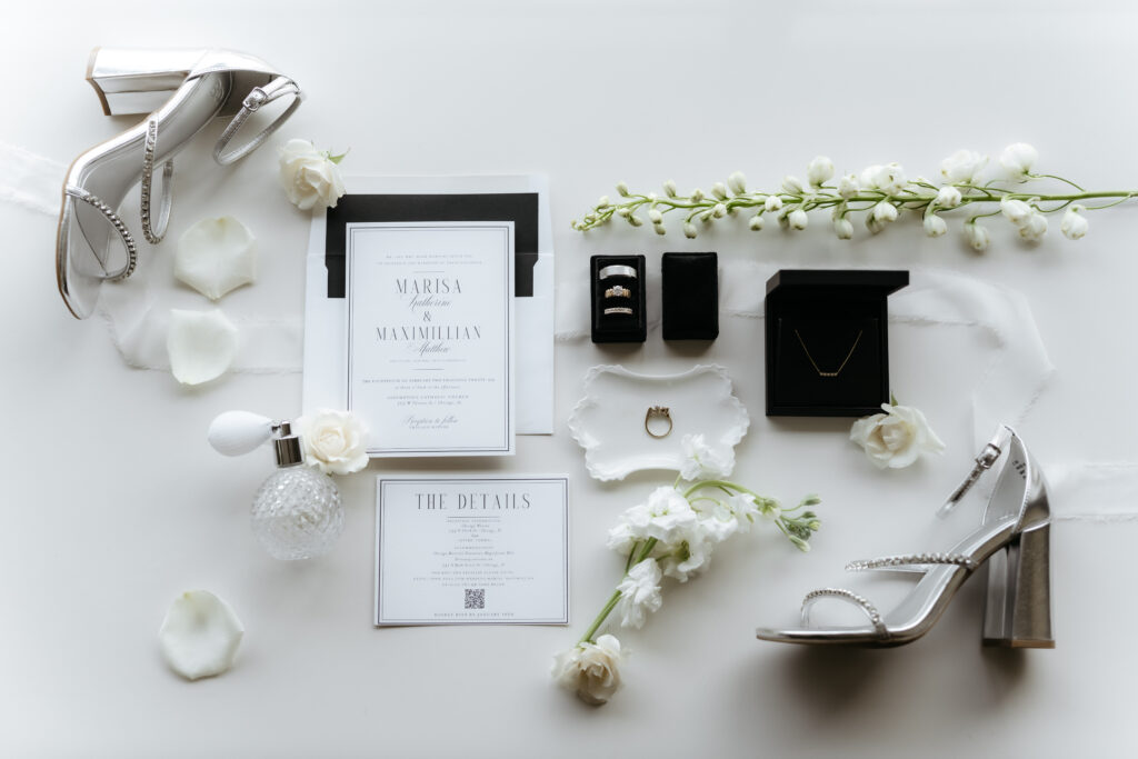 Wedding invitation flat lay featuring silver heels, rings in velvet boxes, a perfume bottle, and white flowers.