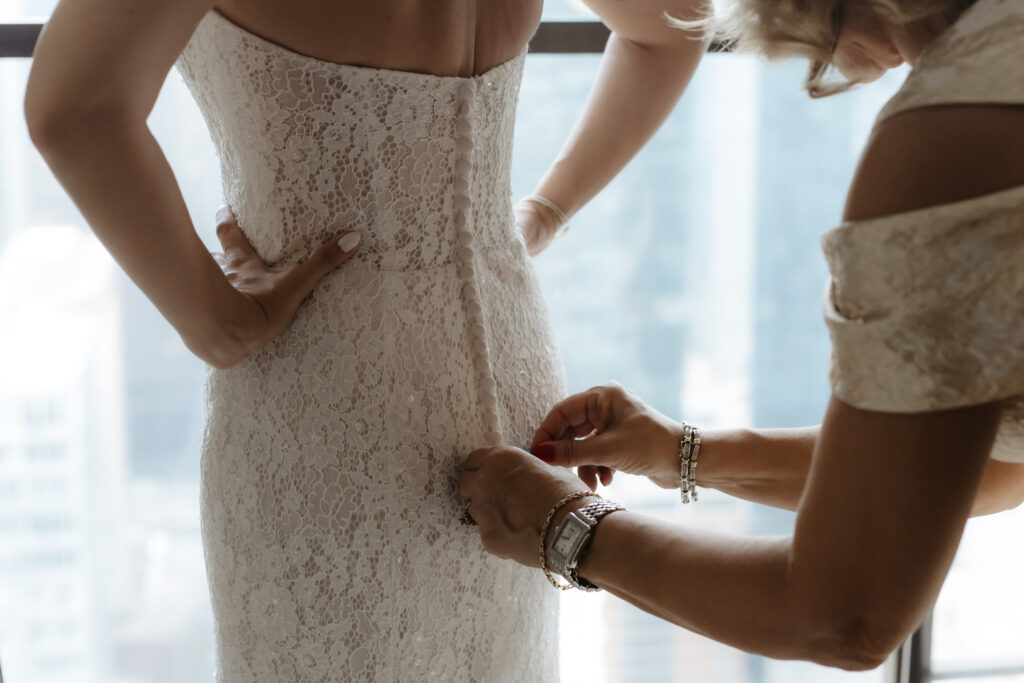 Mother of the bride fastening the back buttons of her daughter's lace wedding gown in front of a city window.